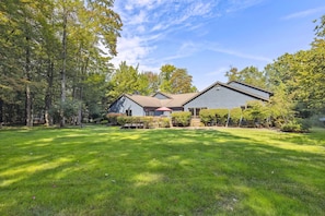 A two-story house with a sloped roof, surrounded by a well-maintained lawn and dense trees.