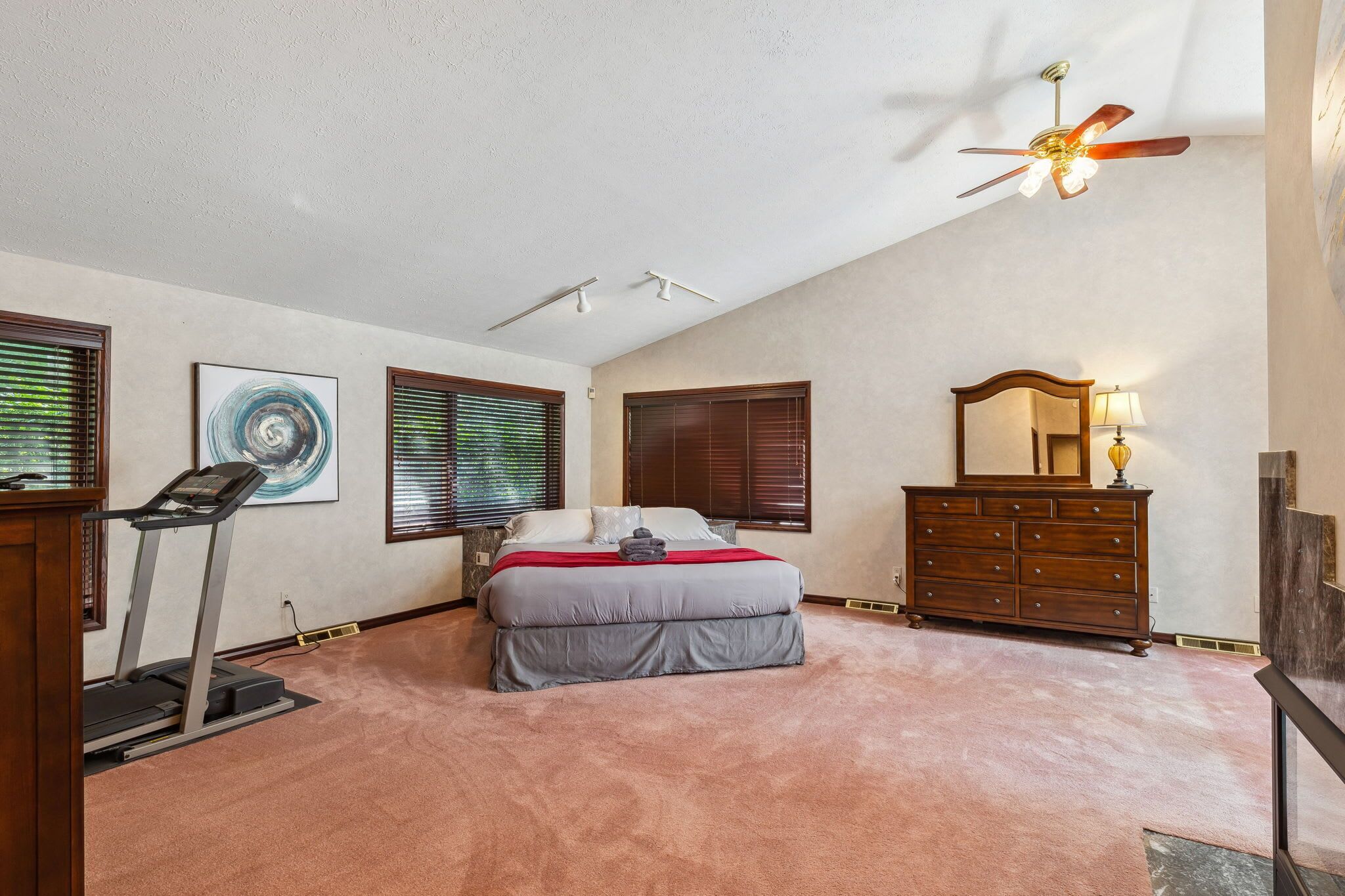 A bedroom with a bed, a treadmill, a dresser, a ceiling fan, and a large window with blinds.