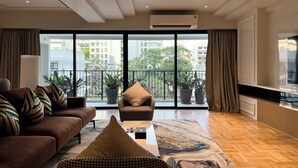 Una sala de estar moderna con sofá, sillón y mesa de centro, ventanas amplias con vistas a edificios y árboles, y un equipo de aire acondicionado en el techo.