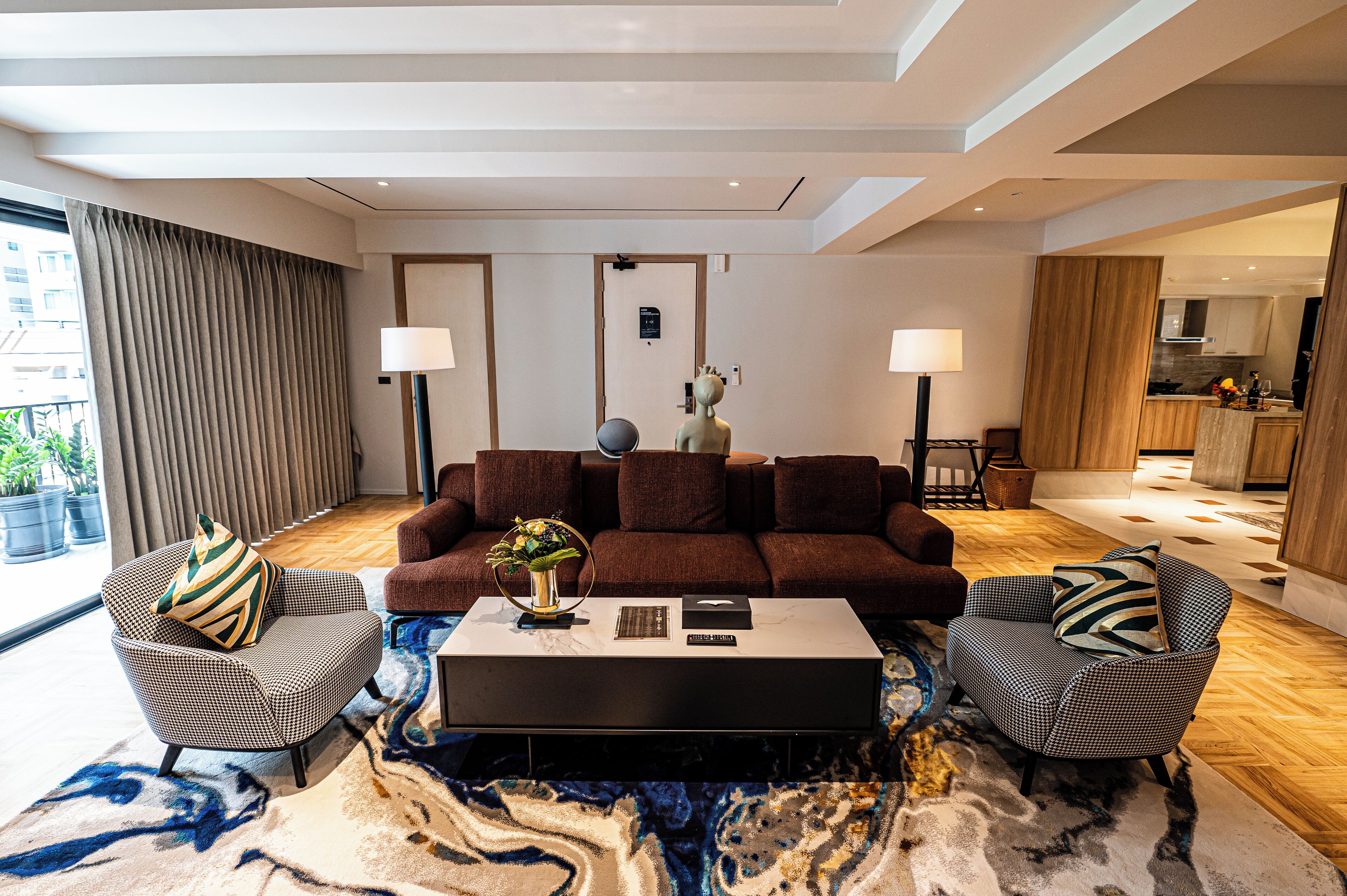 A modern living room with a brown sofa, two patterned armchairs, a coffee table with books and a vase, and a large window with curtains.