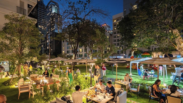 Un área de comedor al aire libre con mesas y sillas, rodeada de vegetación y árboles, con edificios al fondo.