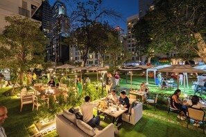 An outdoor dining area with tables and chairs, surrounded by greenery and trees, with buildings in the background. - Floral Court Hotel & Residence Sukhumvit 13 (Bangkok)
