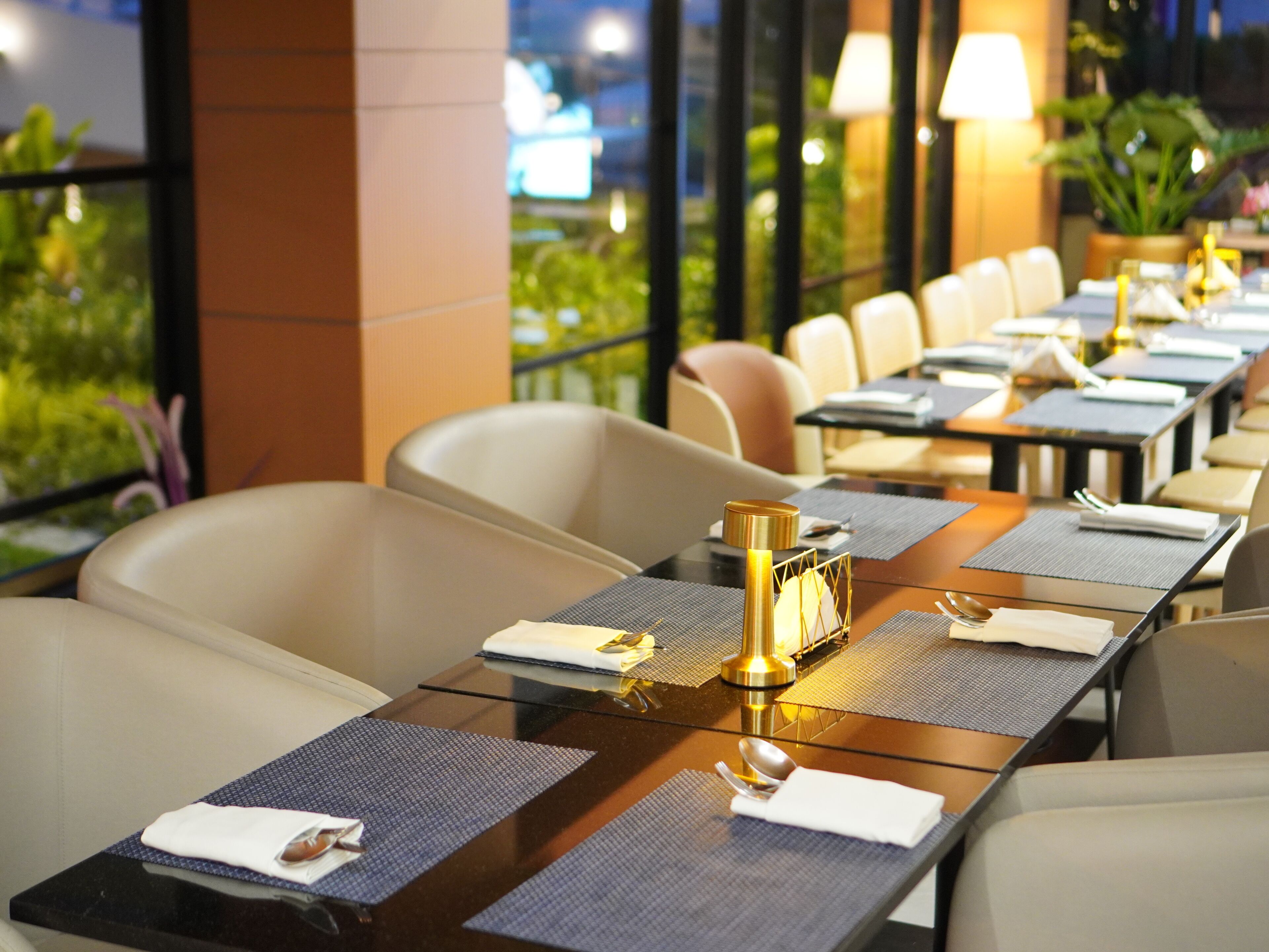Un restaurant où les tables sont dressées pour le service, avec un design moderne, une cloison en verre et des sièges confortables.