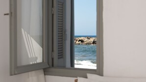Quarto com uma cama, almofada rosa e janela com vista para o mar e rochas.