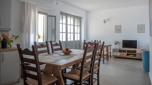 Uma sala de jantar com mesa de madeira, cadeiras e uma fruteira. Há uma televisão e um armário ao fundo.
