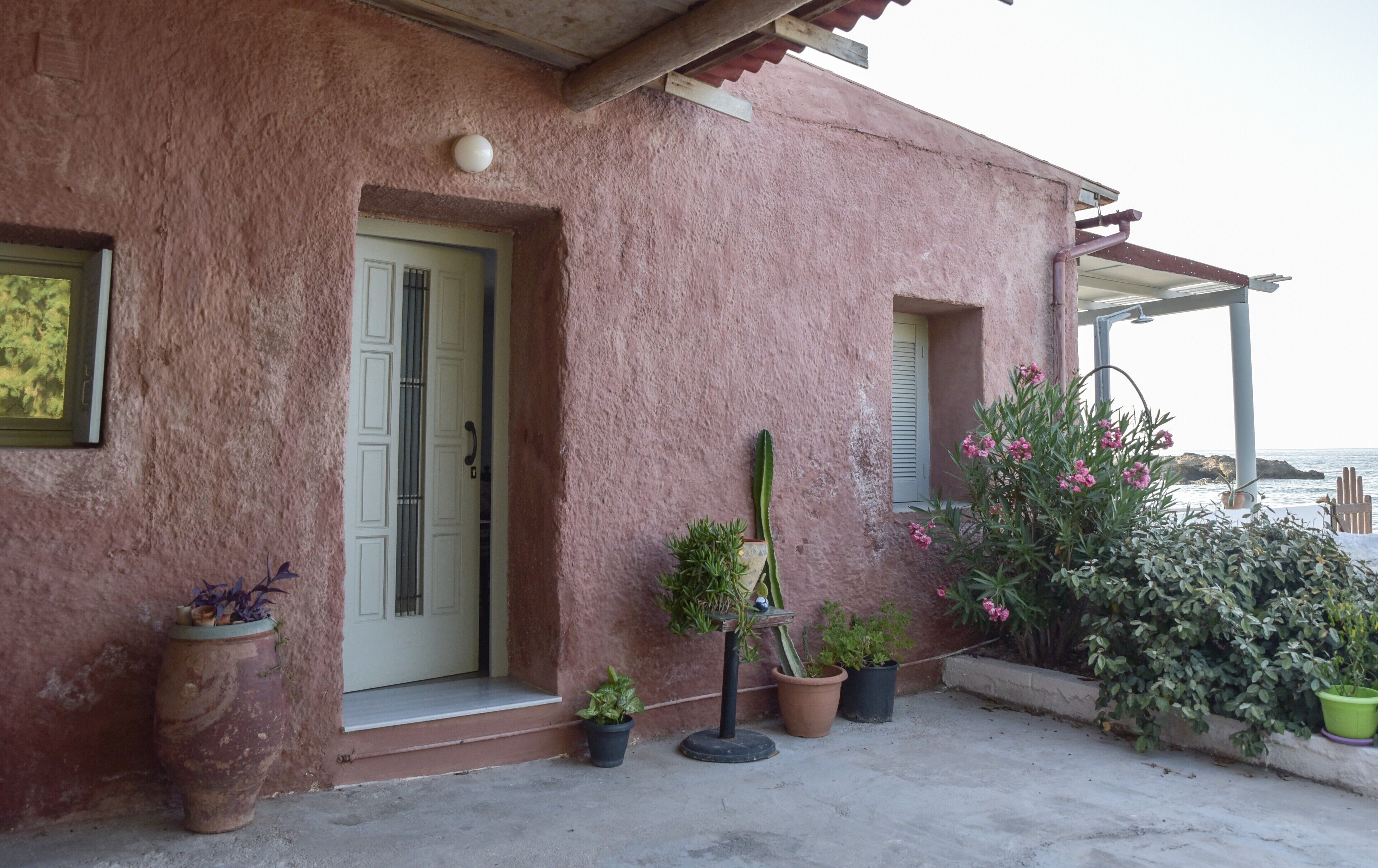 Una casa con fachada de estuco rosado, una puerta blanca y un pequeño porche con plantas en macetas y una silla de metal.
