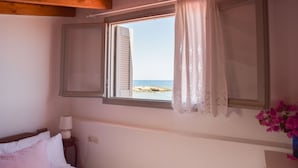 Ein Schlafzimmer mit Blick auf einen Strand, ein Bett mit weißer Bettwäsche, ein Nachttisch mit Lampe und eine Vase mit Blumen.