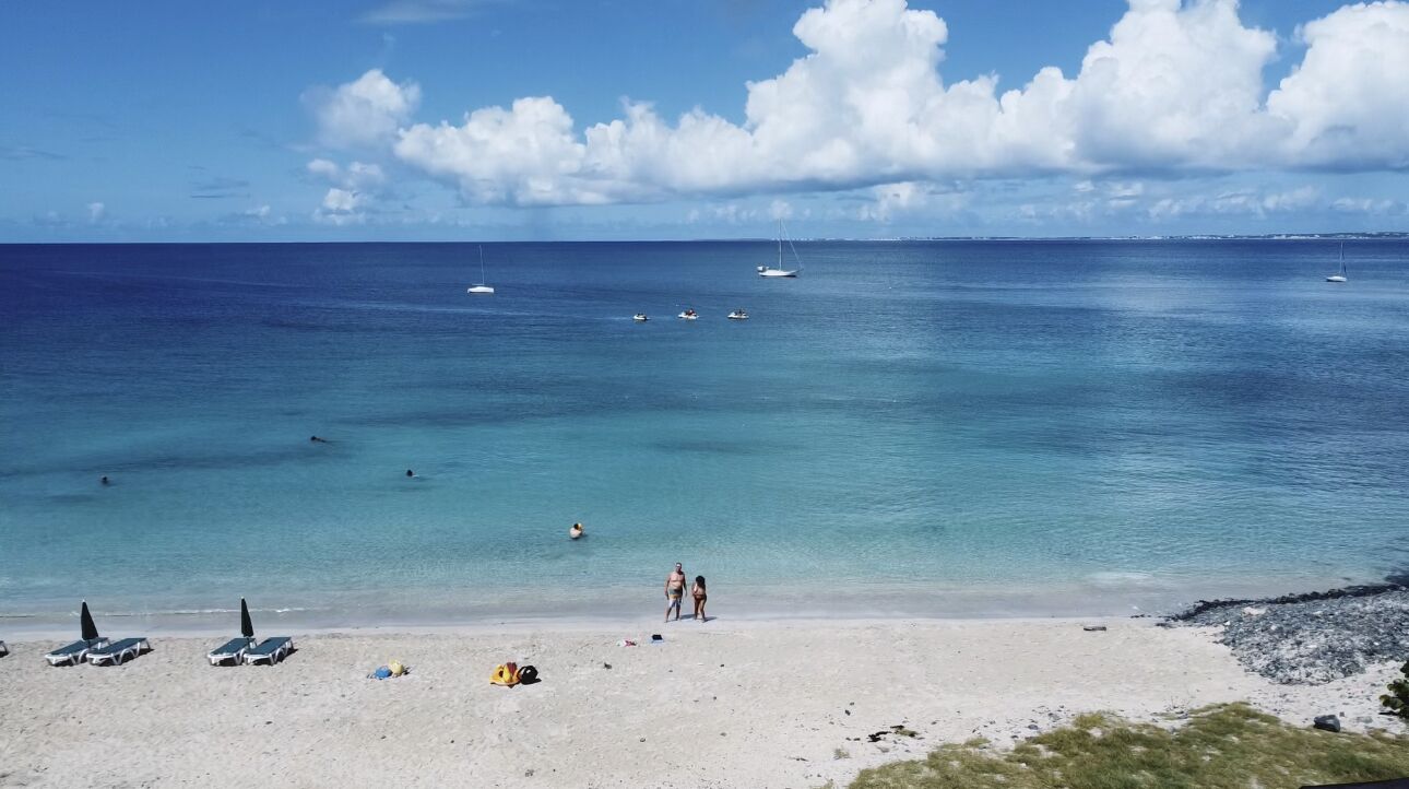 Una playa con aguas turquesas cristalinas, algunas embarcaciones y una orilla arenosa con sombrillas y sillas de playa.