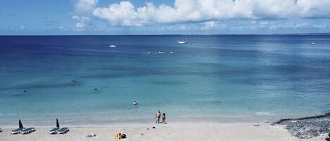 Una playa con aguas turquesas cristalinas, algunas embarcaciones y una orilla arenosa con sombrillas y sillas de playa.