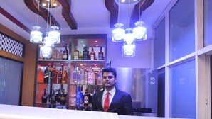 A bar area with a variety of liquor bottles, a man in a suit, and a modern chandelier. - Hotel Aarati (Kathmandu)