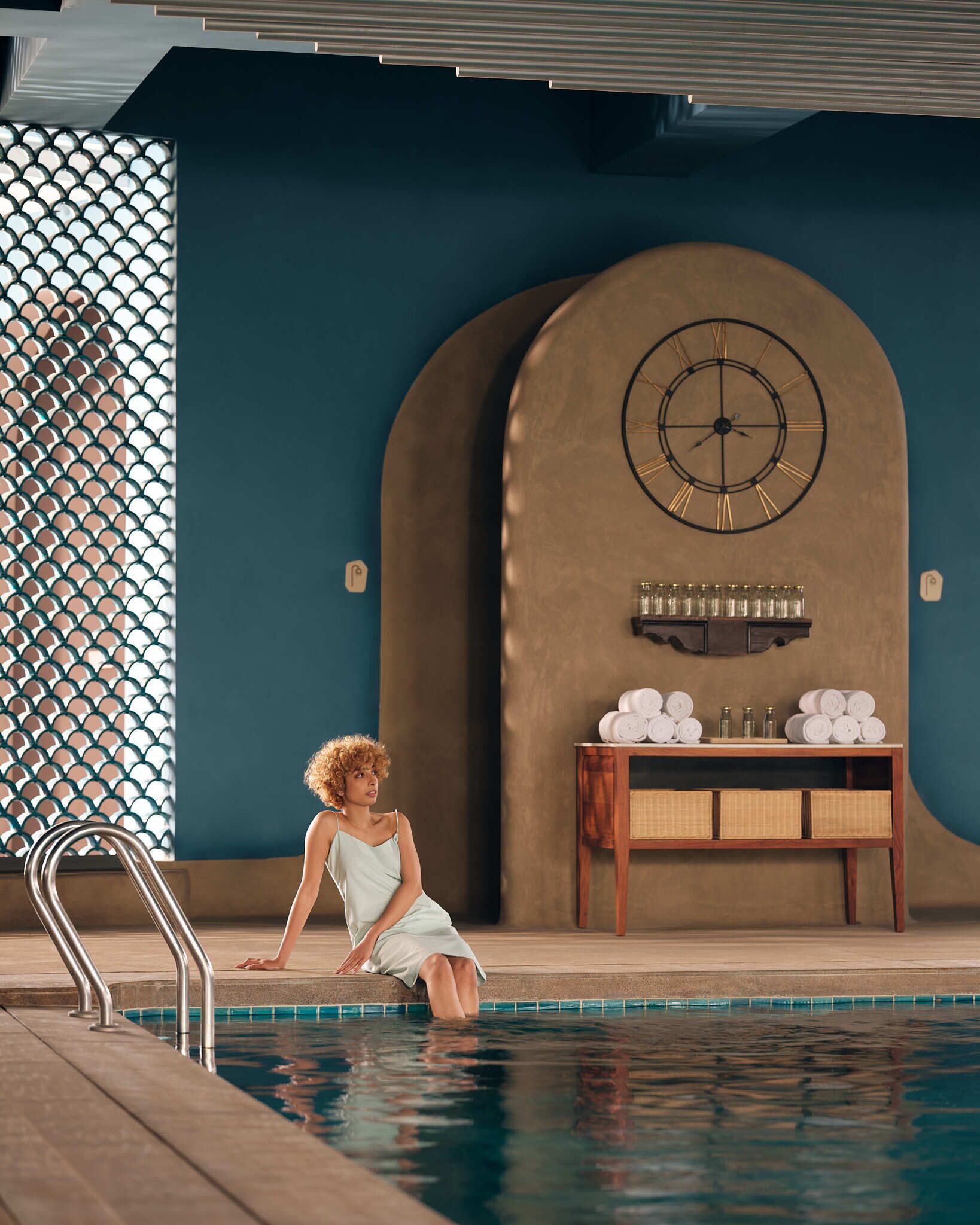 A woman sitting by a pool with a clock and towels on a table in the background.