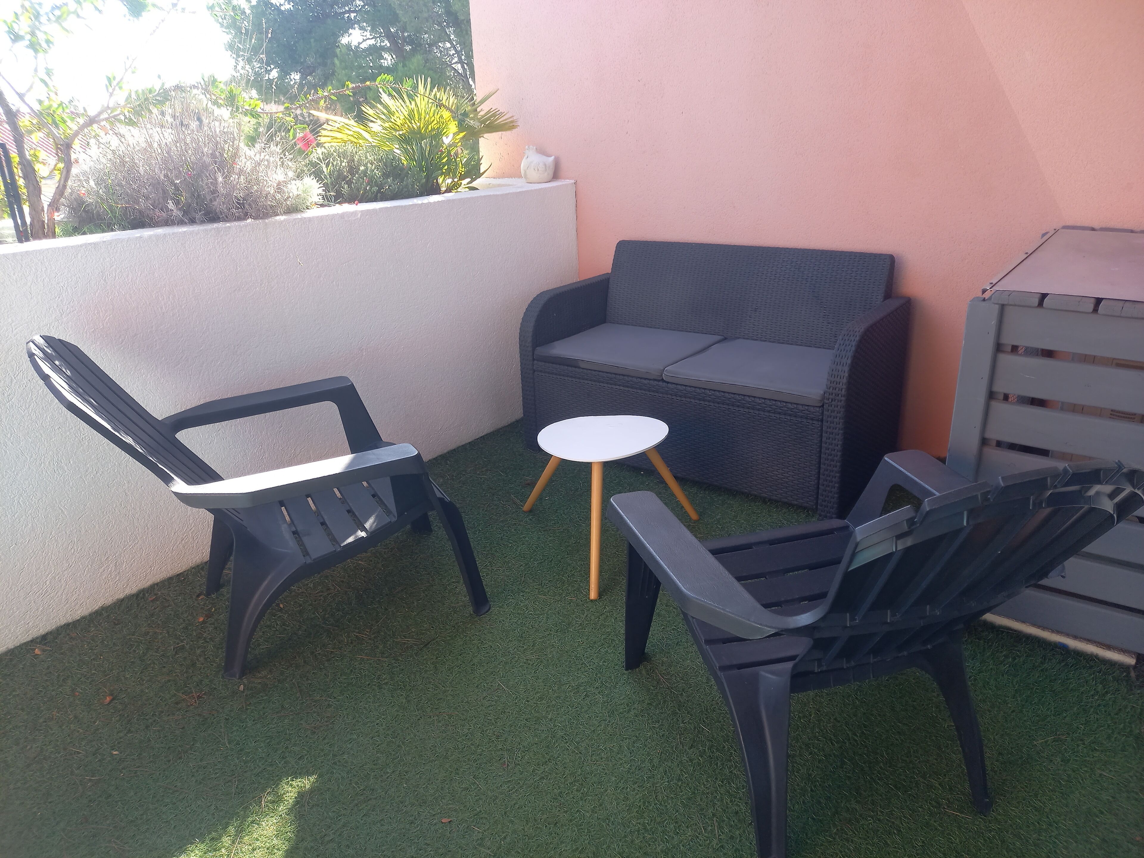 Uma área de pátio com duas cadeiras de plástico preto, um sofá de vime cinza e uma pequena mesa branca com pés de madeira.