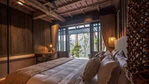 Quarto com cama grande, paredes de madeira, escrivaninha e vista para árvores através de amplas janelas.