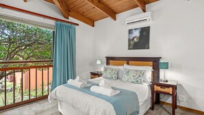 A bedroom with a large bed, a wooden ceiling, an air conditioner, a nightstand, a lamp, and a view of trees through a window. - San Lameer Villa 2704 (Southbroom)