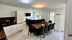 Una cocina moderna con armarios blancos, una mesa de comedor de madera y sillas negras. Hay un televisor montado en la pared y una araña en el techo.