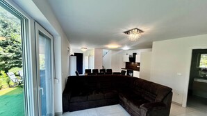 Una sala moderna con un sofá seccional marrón oscuro, una zona de comedor con mesa y sillas, y una cocina con armarios blancos y encimera negra.