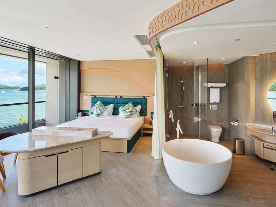 A modern bedroom with a large bed, a sitting area, a bathroom with a bathtub, and a view of the sea.