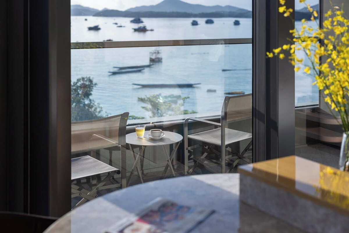Un balcon avec une table et des chaises, un verre à la main, et une vue sur les bateaux qui naviguent.