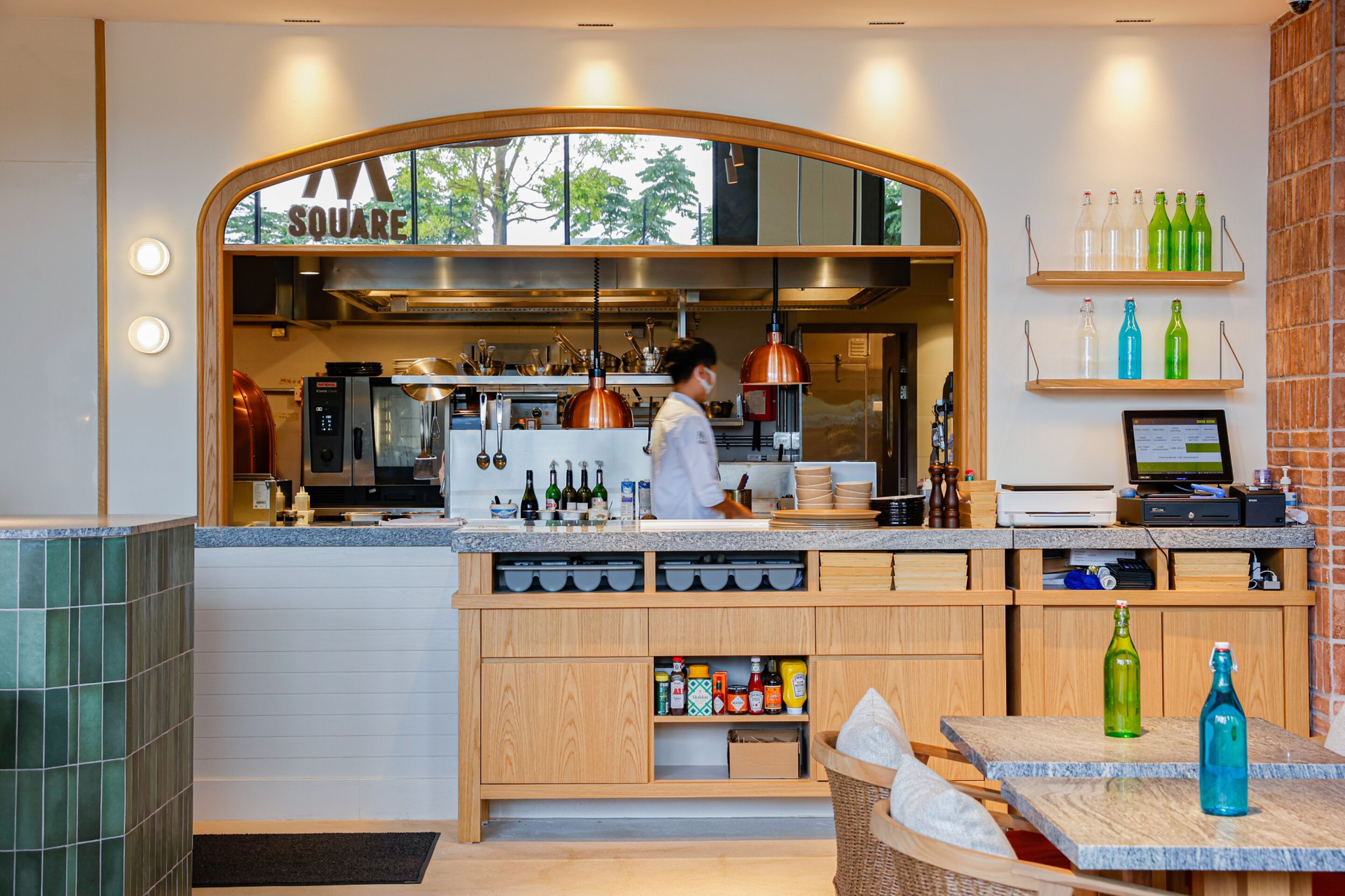 A modern kitchen with a chef preparing food, wooden cabinets, and a bar area with bottles.