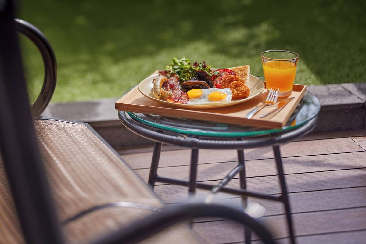 Ein Frühstücksbrett mit Eiern, Speck, Toast und einem Glas Orangensaft auf einem Holztisch im Freien.