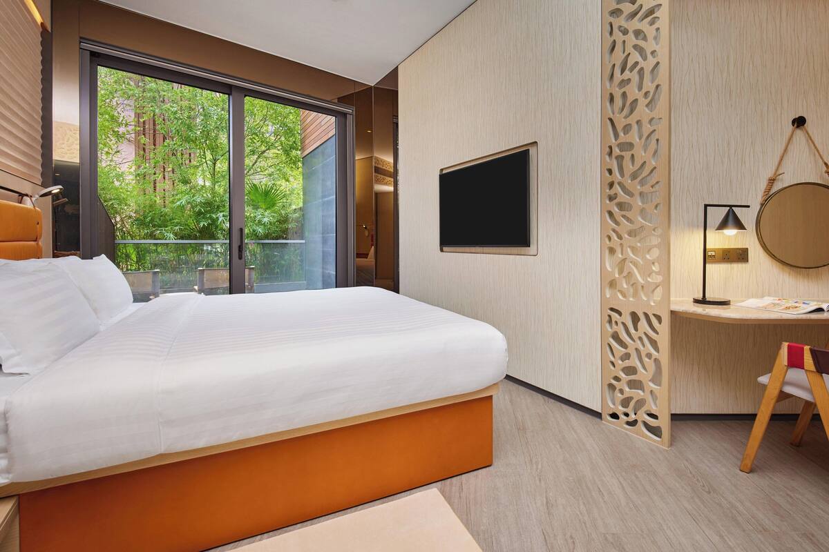 Ein modernes Hotelzimmer mit einem großen Bett, einem Schreibtisch, einem Fernseher und einem Balkon mit Blick ins Grüne.