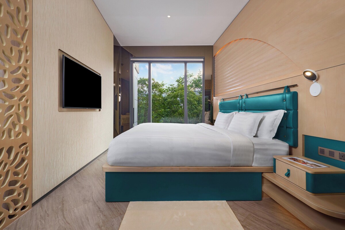 Une chambre moderne avec un grand lit, un téléviseur fixé au mur et une vue sur la verdure par la fenêtre.
