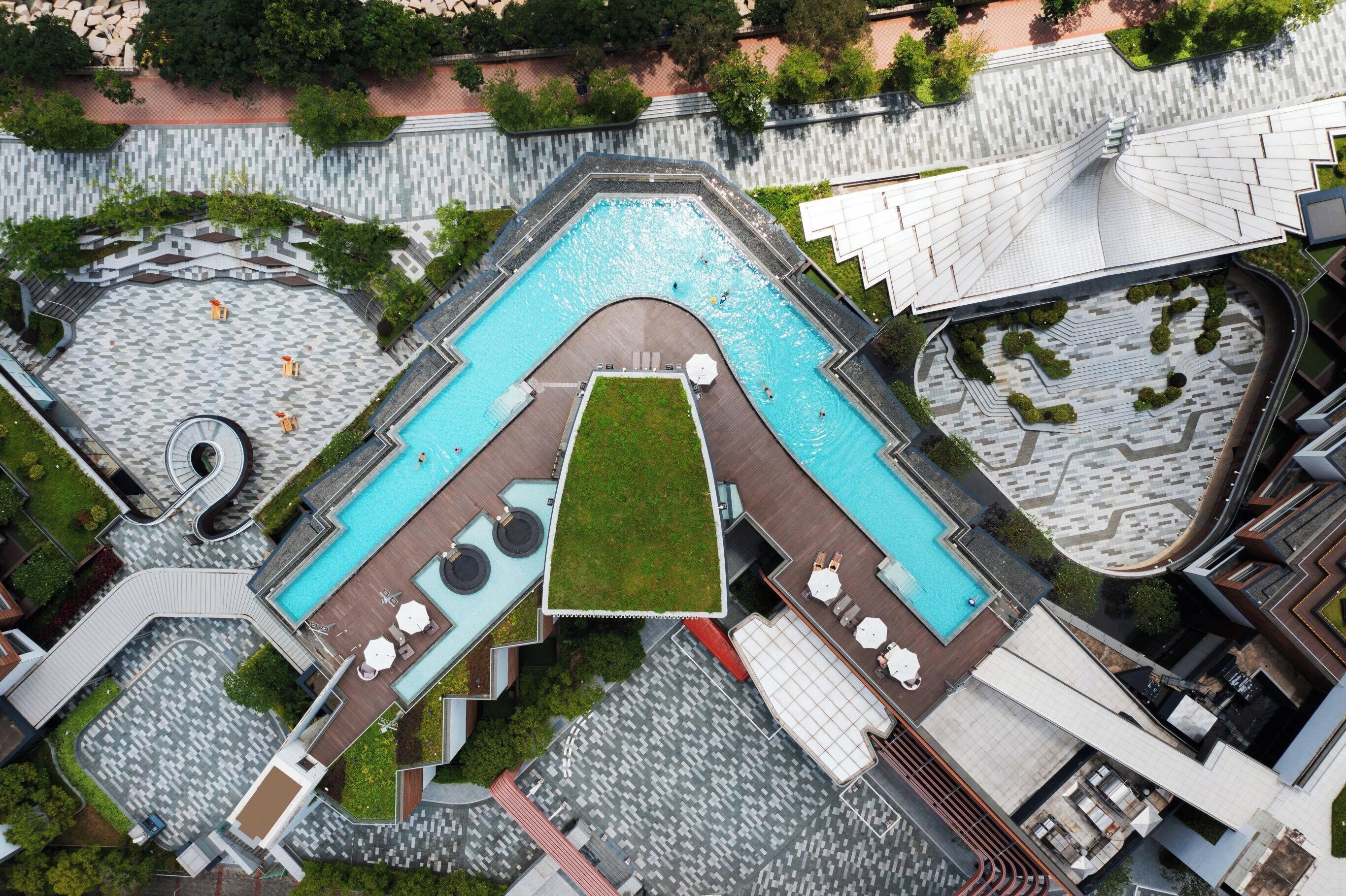 An aerial view of a modern hotel with a central swimming pool, multiple lounge areas, and a landscaped garden.
