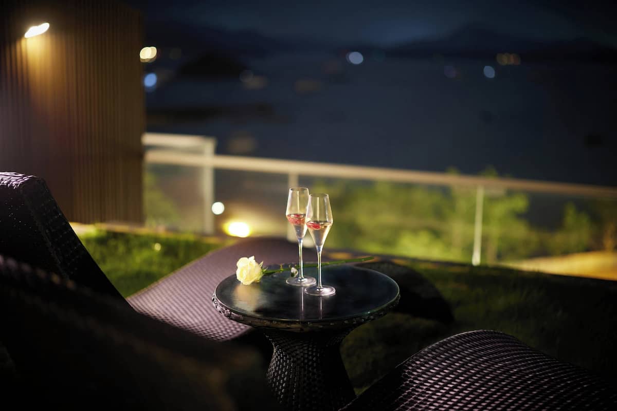 Ein Tisch mit zwei Weingläsern und einer weißen Rose, vor einer nächtlichen Kulisse mit Blick auf Grünflächen und entfernte Lichter.