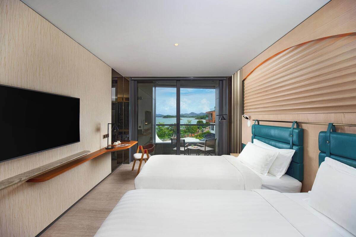 Ein modernes Hotelzimmer mit einem großen Flachbildfernseher, zwei Betten mit Türkis-Kopfteilen, einem Schreibtisch mit Stuhl und einem Balkon mit Bergblick.