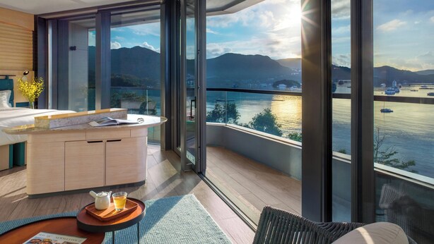 Una habitación de hotel moderna con un balcón grande, una cama, una mesita con bandeja y vistas a una masa de agua con barcos.