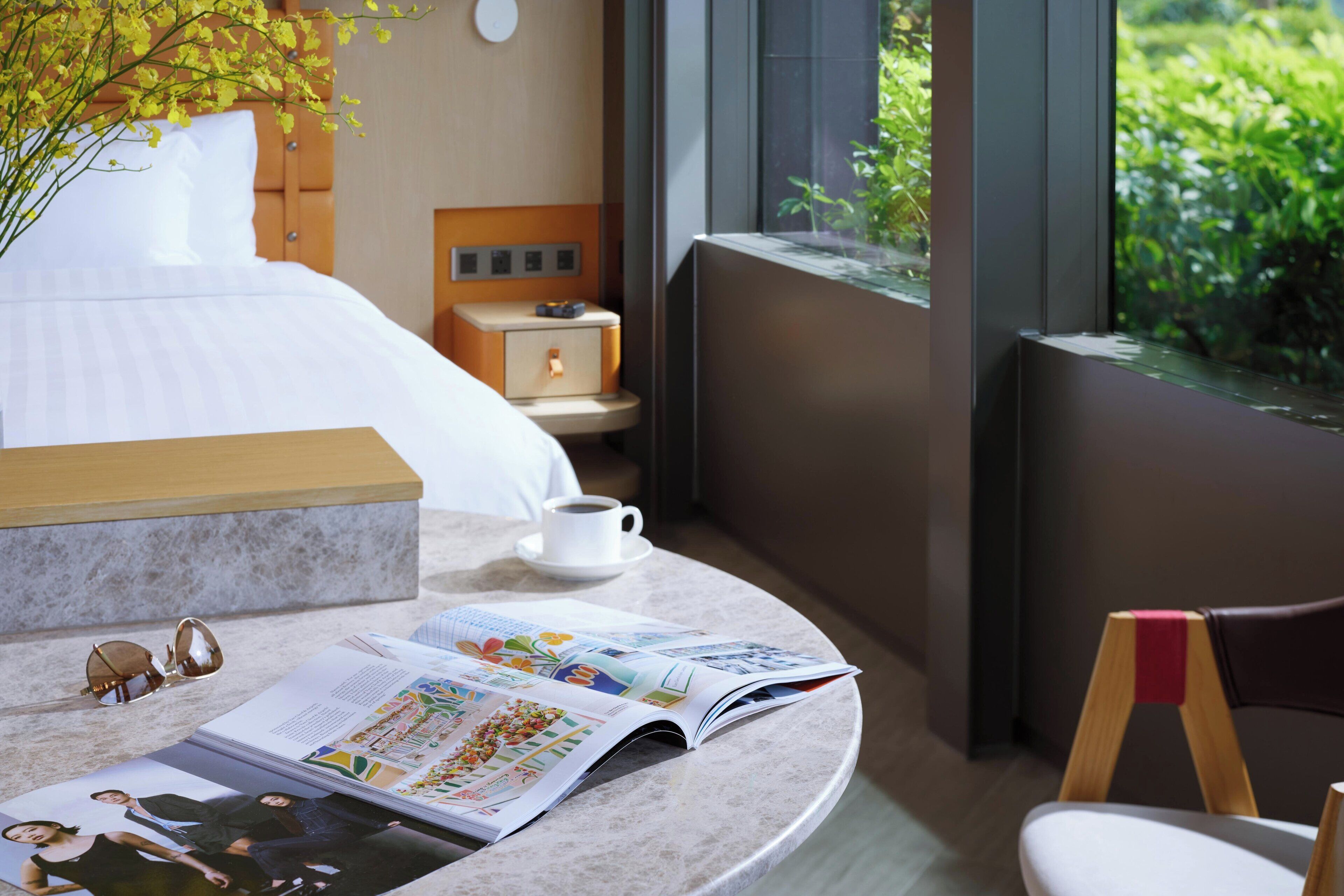 A modern hotel room with a bed, a bedside table with a cup, a magazine, and a window with a view of greenery.