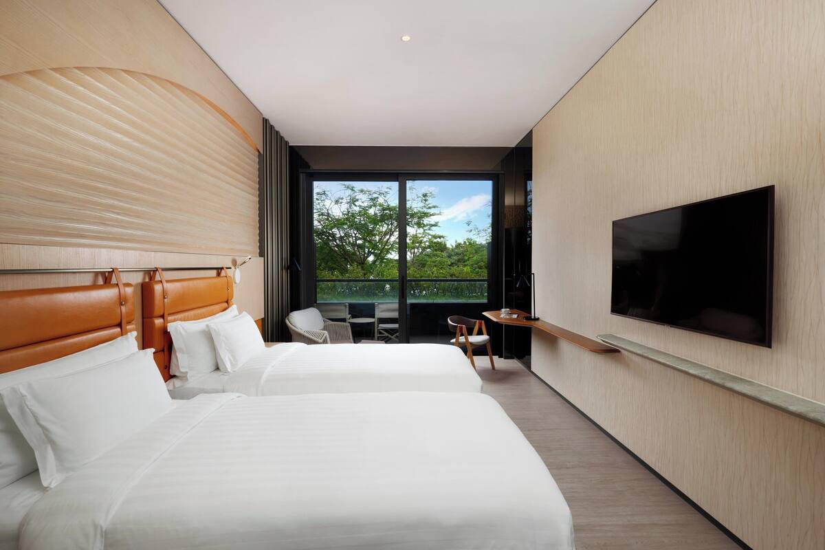 Ein modernes Hotelzimmer mit zwei Betten, einem Fernseher und einem Balkon mit Blick auf Bäume.