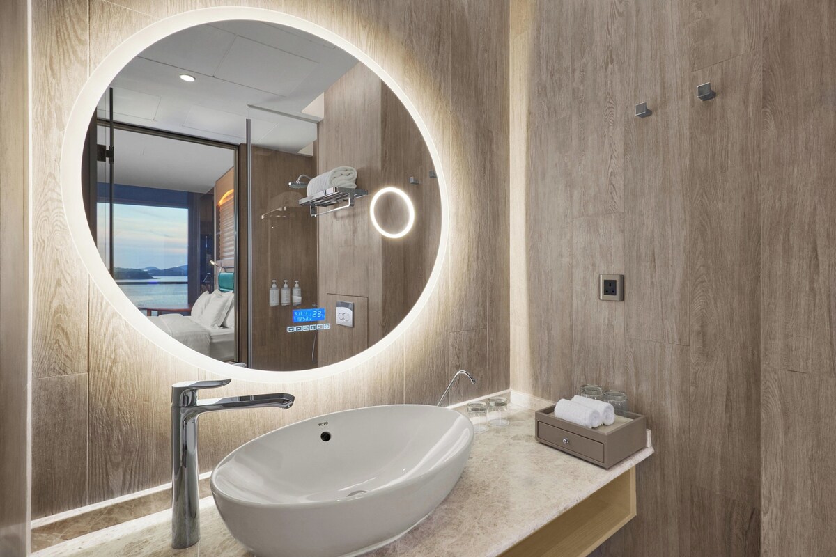 Une salle de bain moderne avec un grand miroir ovale, un lavabo sur colonne blanc et un meuble-lavabo en bois.