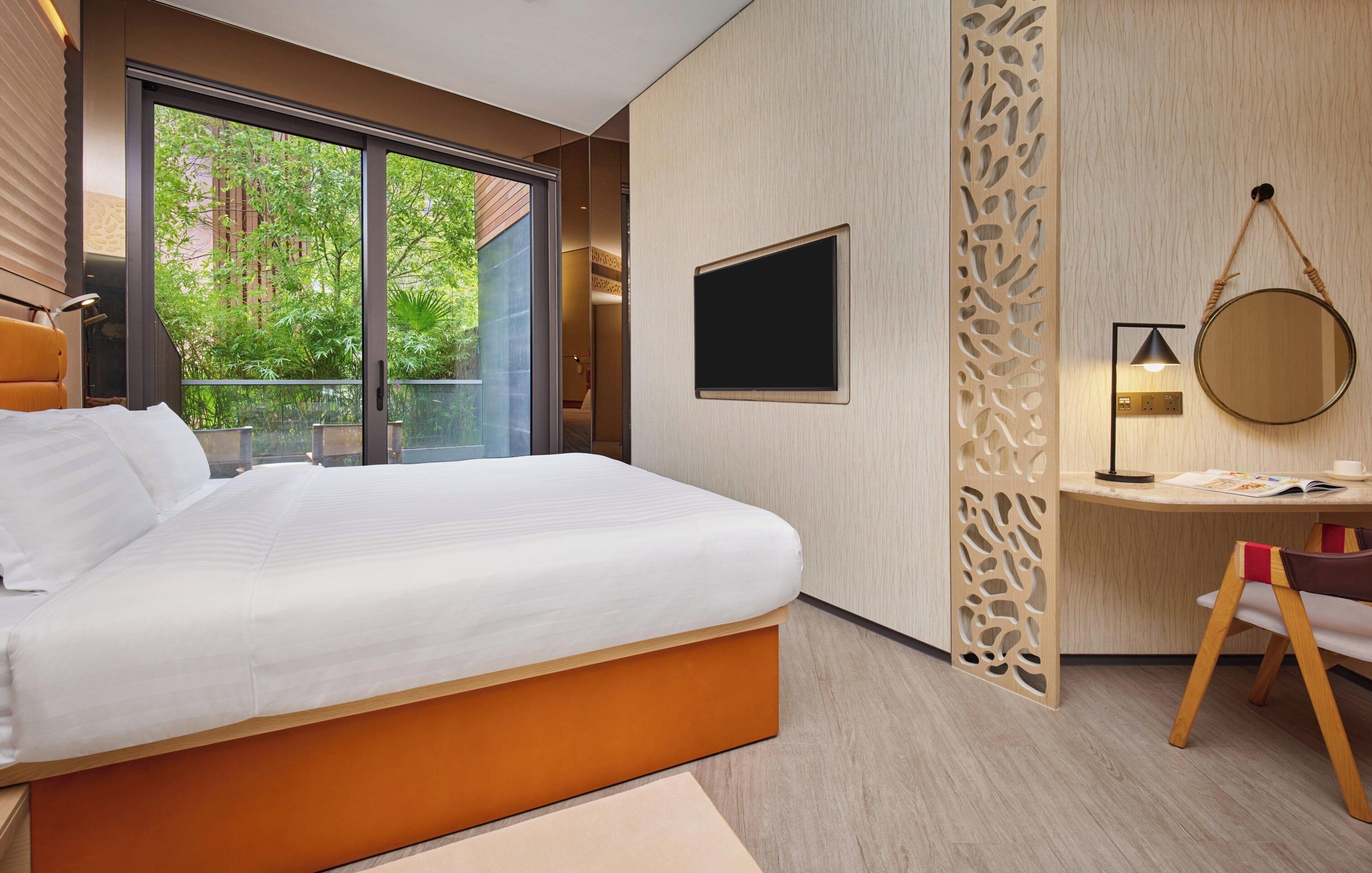 A modern hotel room with a large bed, a desk, a TV, and a balcony with a view of greenery.