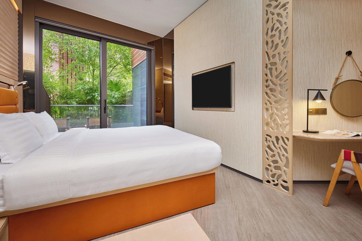 Une chambre d’hôtel moderne dotée d’un grand lit, d’un bureau, d’une télévision et d’un balcon offrant une vue sur la verdure.