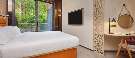A modern hotel room with a large bed, a desk, a TV, and a balcony with a view of greenery.