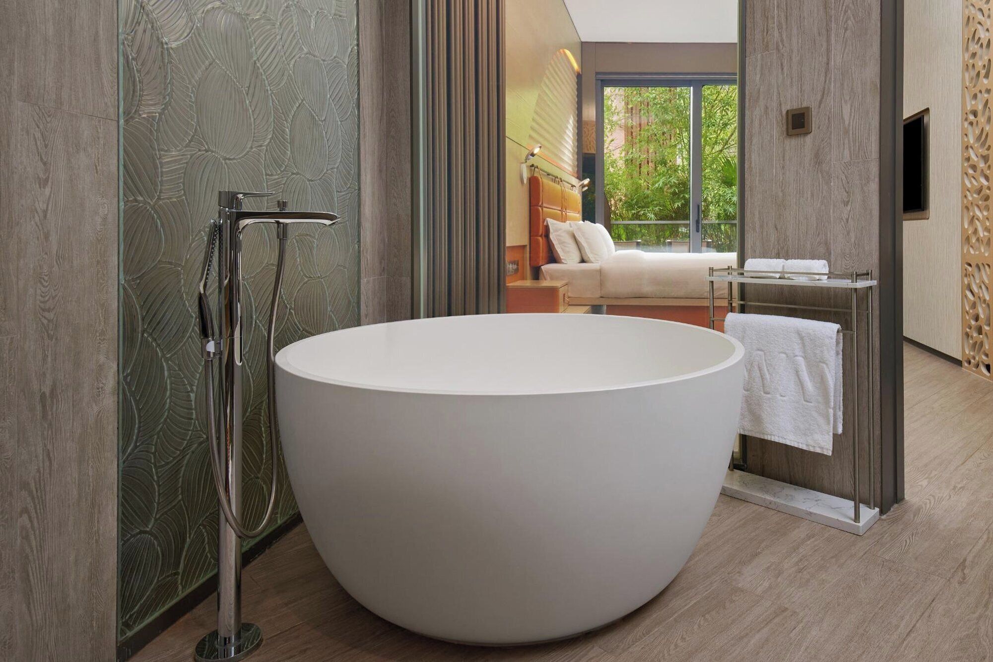 A modern bathroom with a large freestanding bathtub, a glass shower enclosure, and a view of a balcony with a sofa and a TV.