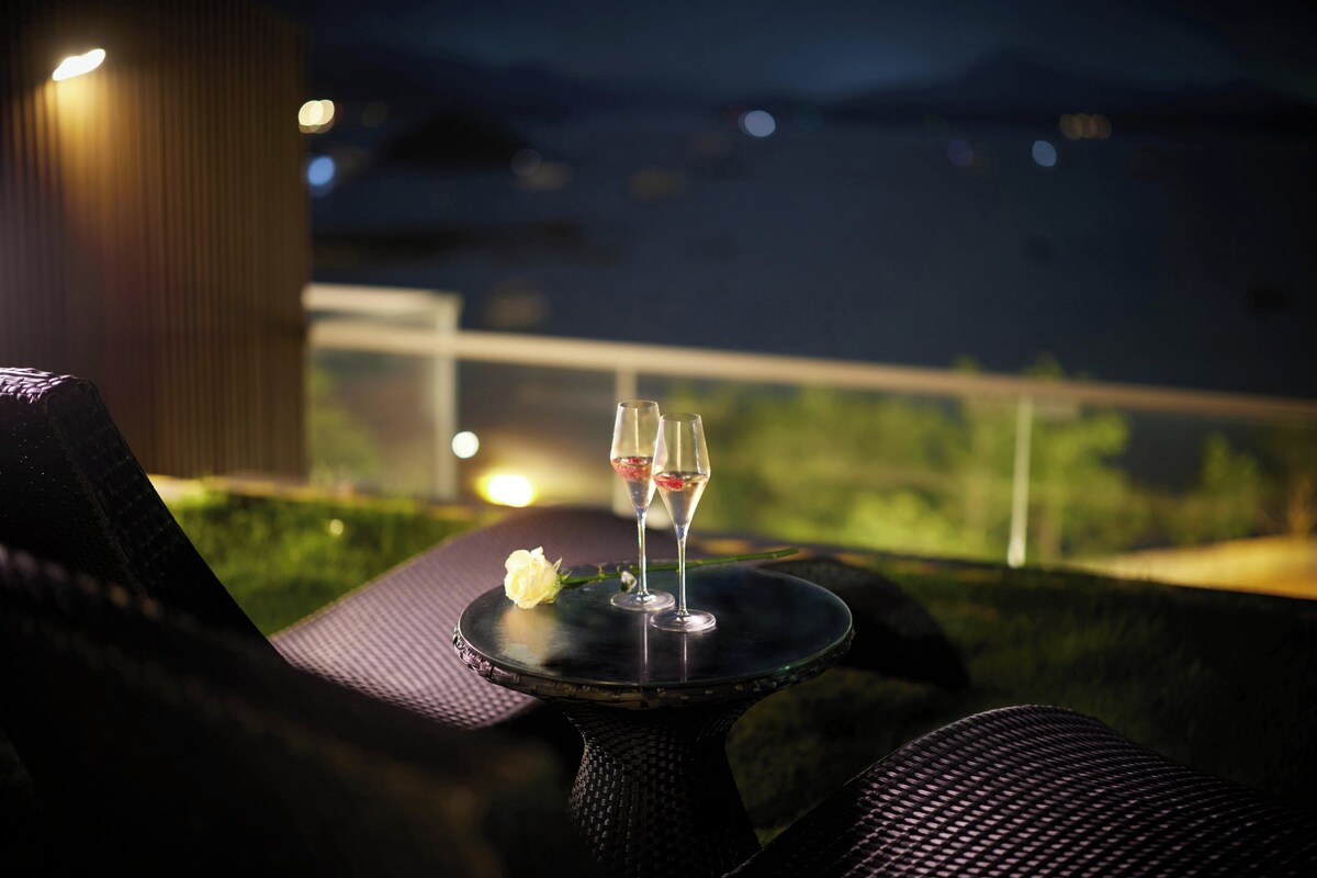 Une table avec deux verres de vin rouge et une rose blanche, placée sur un fond nocturne offrant une vue sur la verdure et des lumières lointaines.