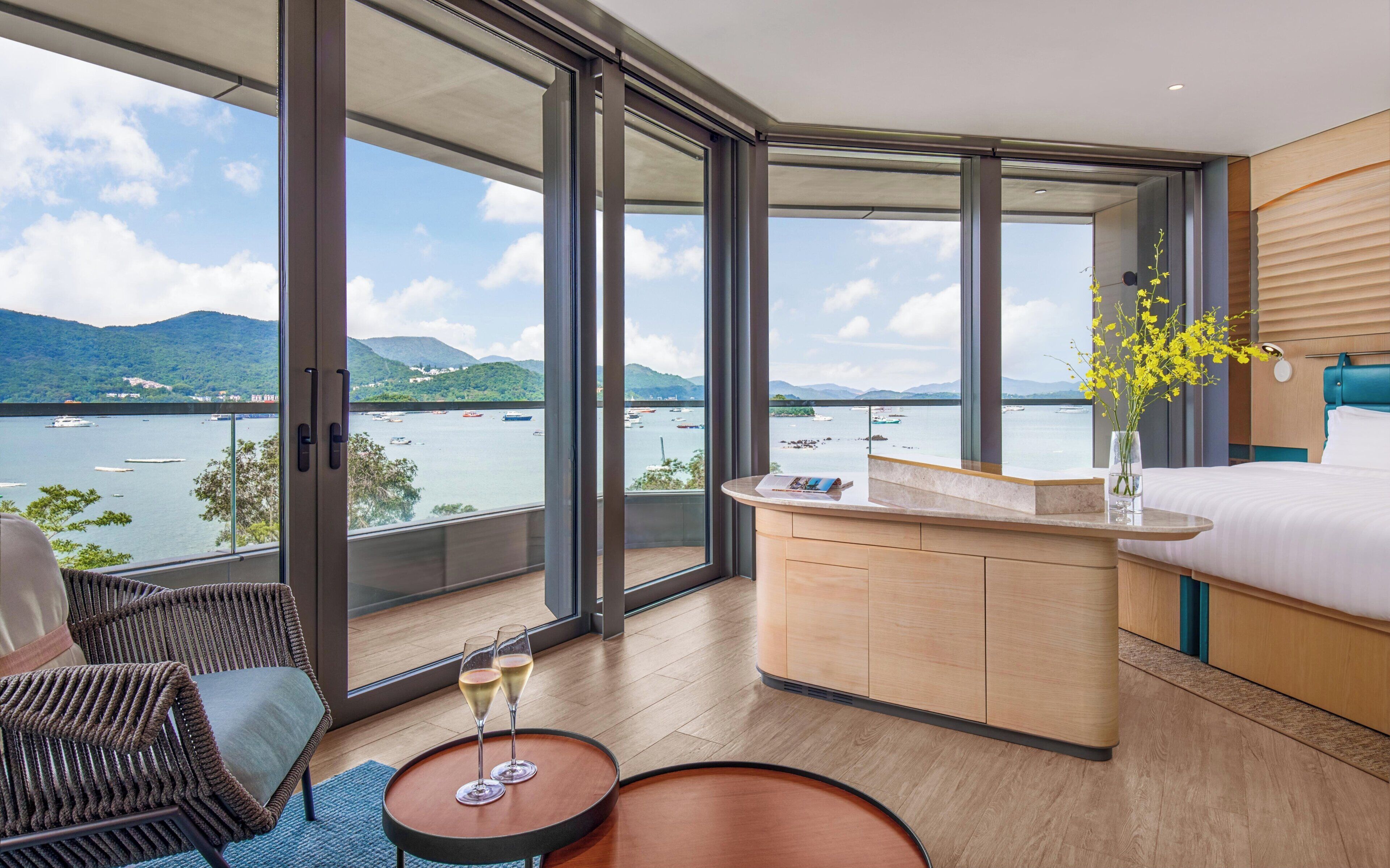 A modern hotel room with a large balcony, a bed, a small desk, and two glasses of champagne on a round table.
