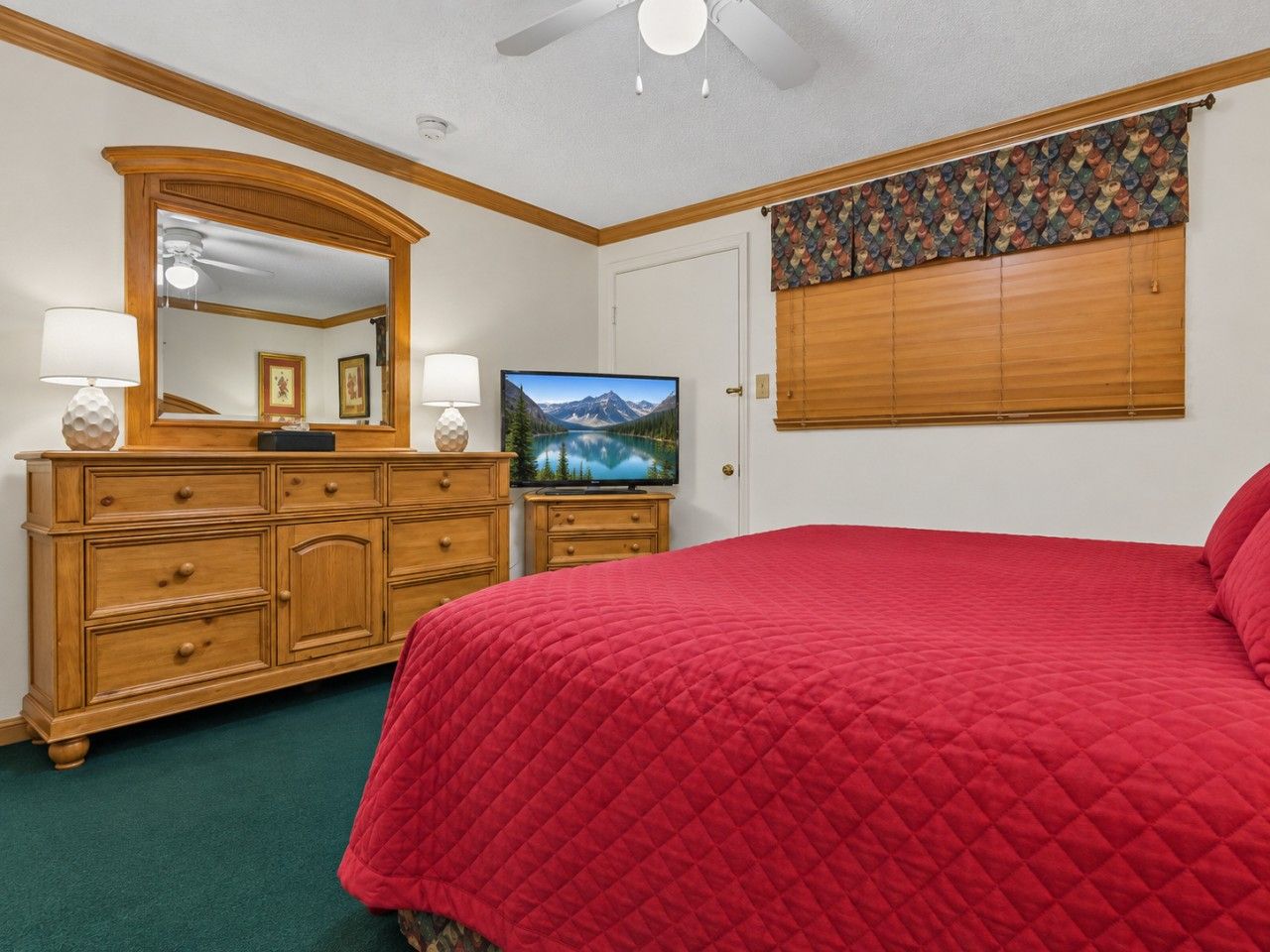 Un dormitorio con colcha de patchwork rojo, un aparador de madera con cajones, un televisor sobre un soporte y un ventilador de techo.