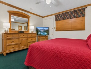 A bedroom with a red quilted bedspread, a wooden dresser with drawers, a television on a stand, and a ceiling fan. - Beautiful Views New Deck Wi-Fi (Snowshoe)