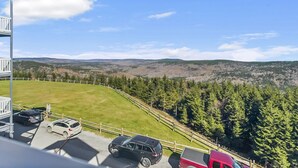 Vista de uma varanda com vista para um estacionamento com vários carros, um campo gramado e uma encosta arborizada.