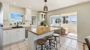 Une cuisine spacieuse avec des meubles blancs, un îlot central en bois doté de tabourets, et de grandes fenêtres offrant une vue sur l’extérieur.