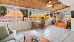 Une pièce spacieuse avec un plafond en bois, de grandes fenêtres offrant une vue sur le coucher de soleil, un coin repas avec des chaises, un canapé et un escalier menant à un niveau supérieur.