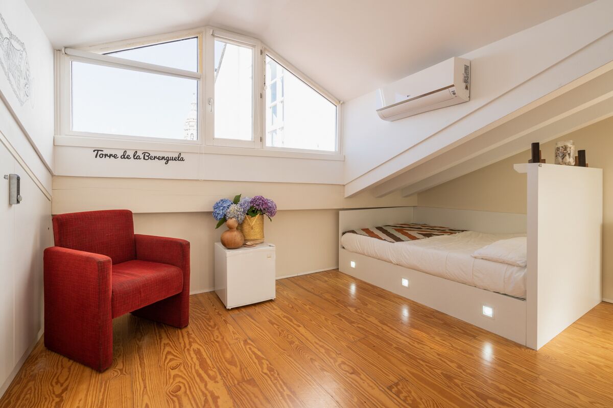 Un dormitorio pequeño y moderno con suelo de madera, una silla roja, una cama blanca y una ventana con vista a un edificio.