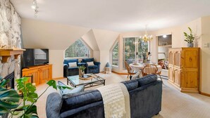 A spacious living room with a fireplace, a sofa, a television, and a dining area with a chandelier. - 31GlenEagle Cozy Townhome | 2 Bedroom 2 Bathroom, GlenEagle Townhome Whistler, Golf/Ski community (Whistler)