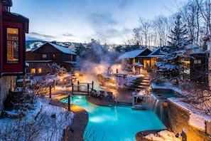 Un complejo invernal con piscina central, cabañas de madera y un paisaje nevado.