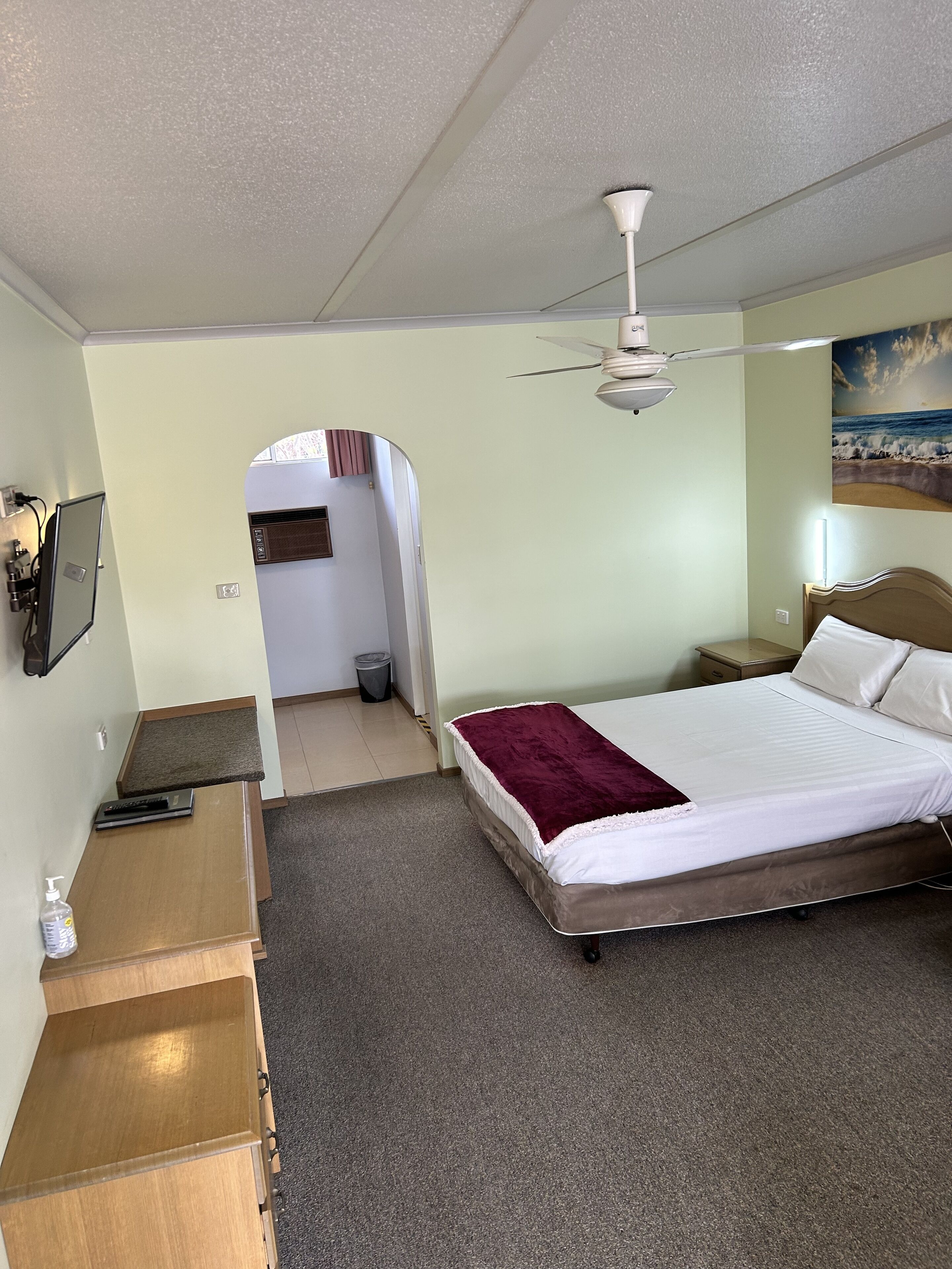 Habitación de hotel con cama, escritorio, ventilador de techo, un cuadro y vista al pasillo.