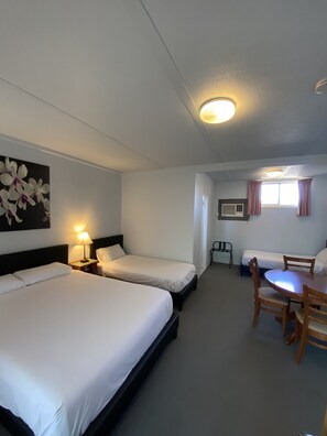 A hotel room with two single beds, a dining table with chairs, a small table with a chair, a wall-mounted air conditioner, and a window with curtains.