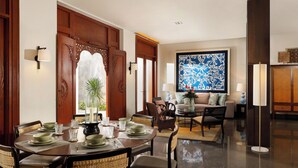 Uma sala de jantar com uma mesa redonda posta para seis pessoas, rodeada por cadeiras de vime. Uma janela grande com vista para o verde é visível ao fundo.